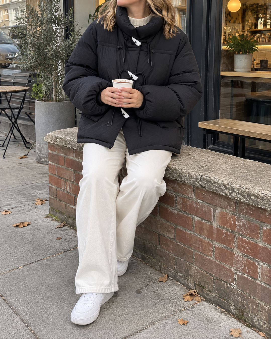 Ombeline - Toggle Button Puffer Jacket in black, on a low wall outside a café on a grey afternoon