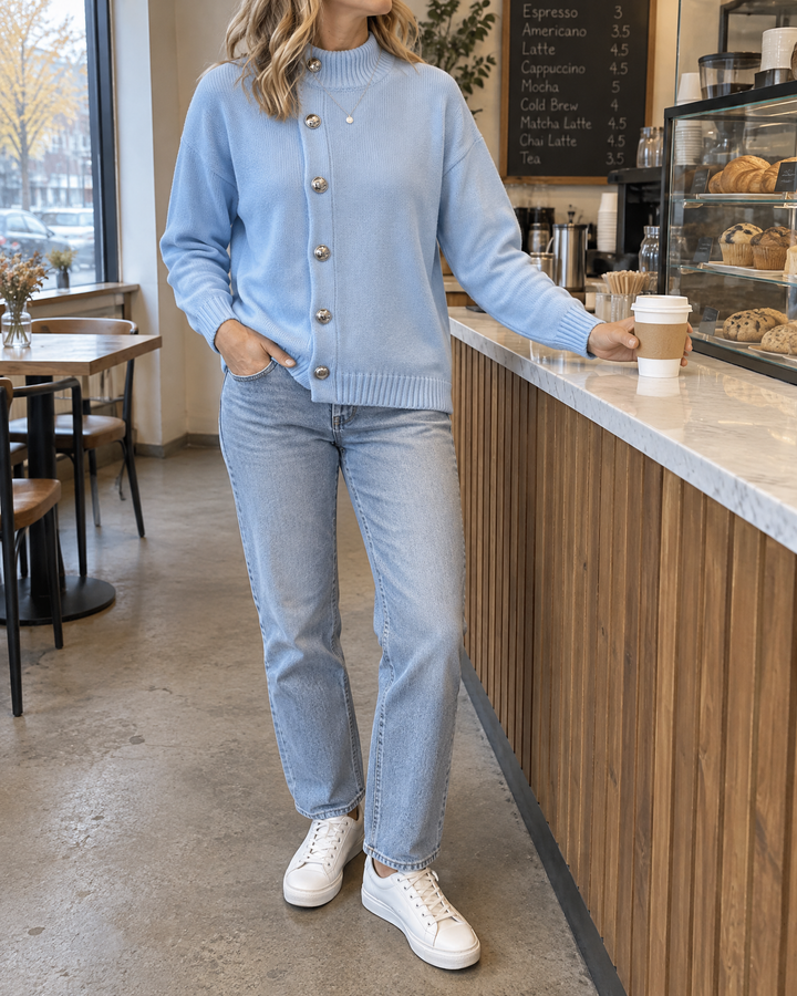 Sixtine - Silver Button Mock Neck Sweater in sky blue, at a café counter on an overcast morning